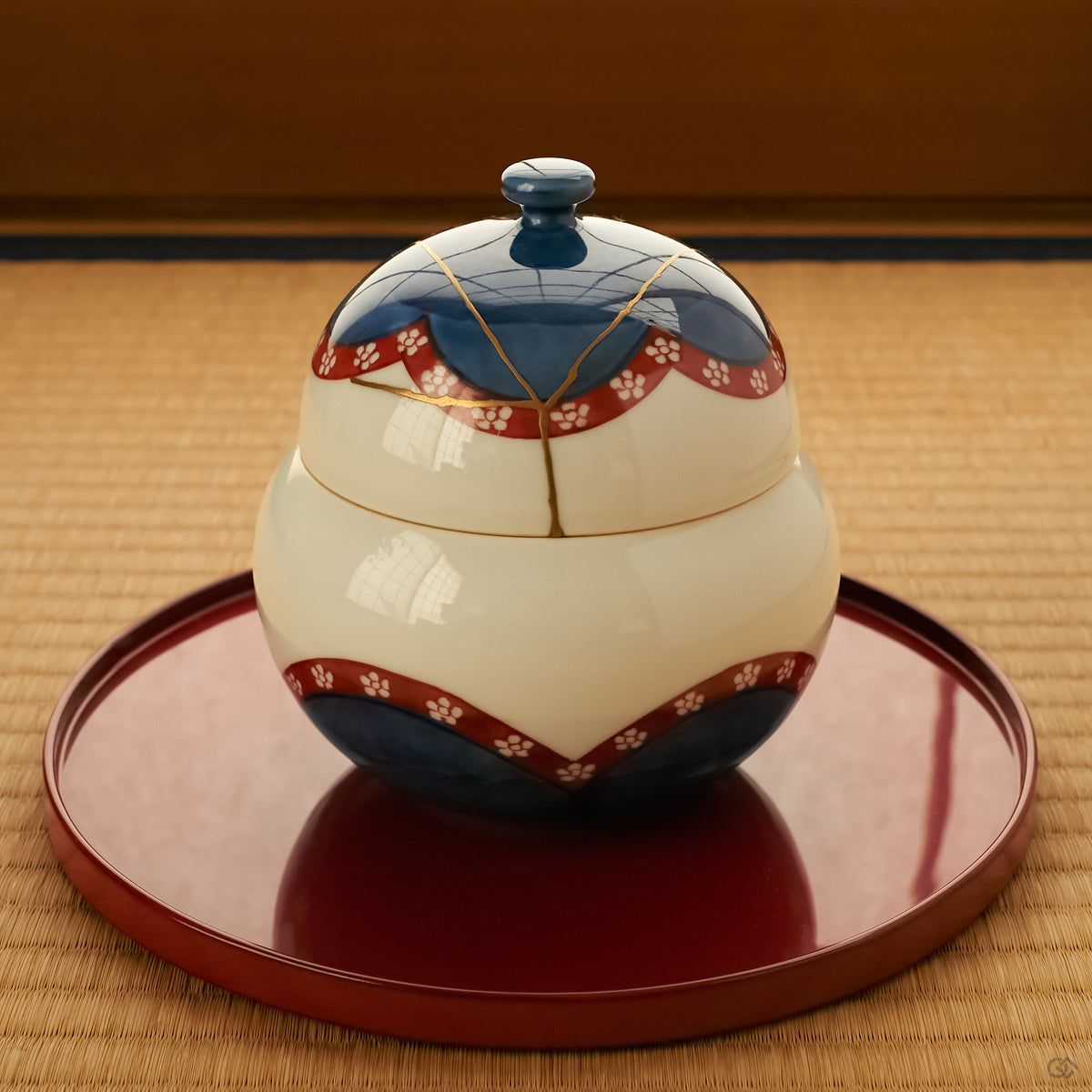 Arita ware lidded bowl with cobalt-blue and vermilion plum-blossom decoration and a 24K gold kintsugi lid, set on a round vermilion lacquer tray over a tatami mat.