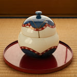 Arita ware lidded bowl with cobalt-blue and vermilion plum-blossom decoration and a 24K gold kintsugi lid, set on a round vermilion lacquer tray over a tatami mat.