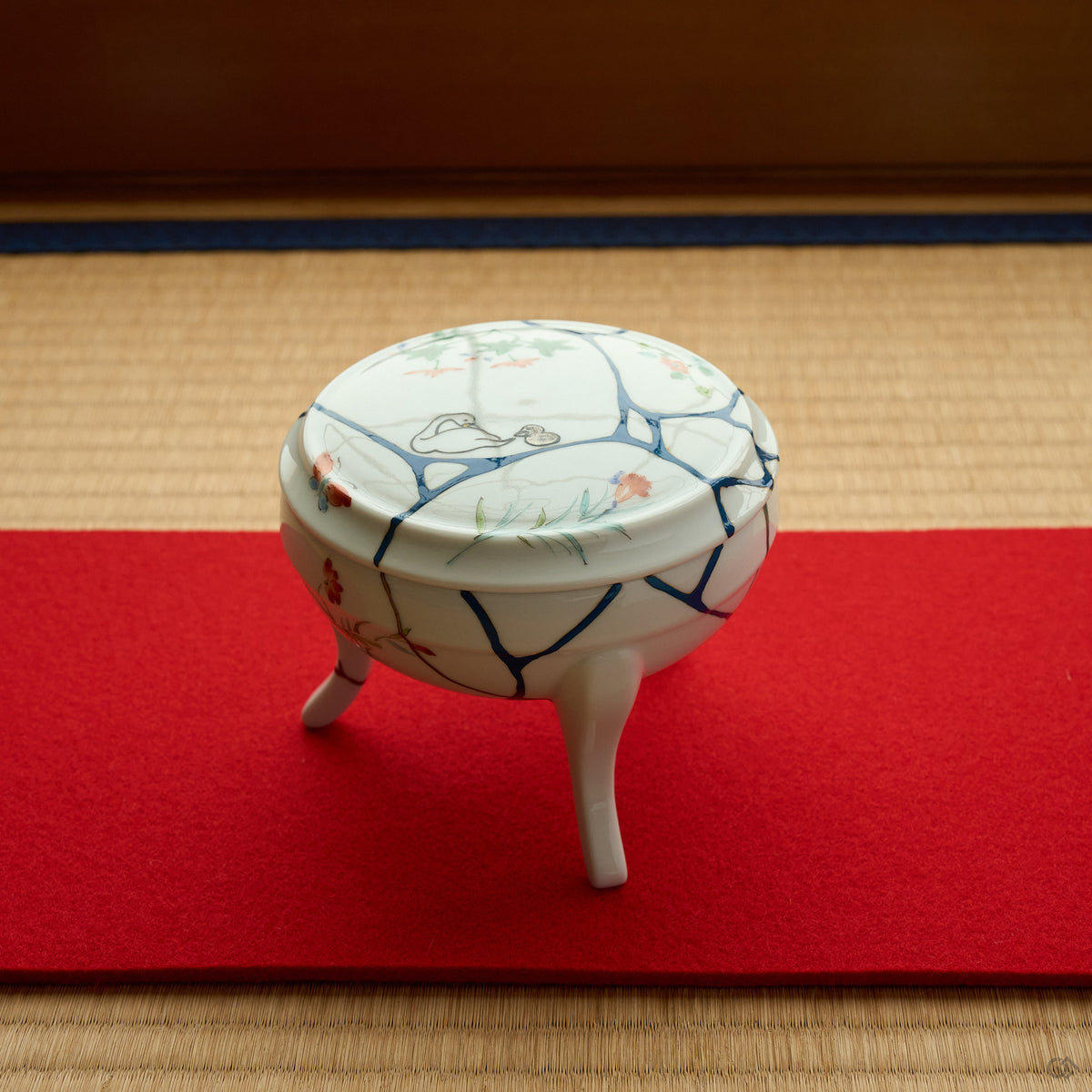 Kintsugi Arita lidded bowl set on a red felt runner over tatami, white porcelain with blue urushi lines and swan maki-e on the lid, 24K gold seams catching the light.