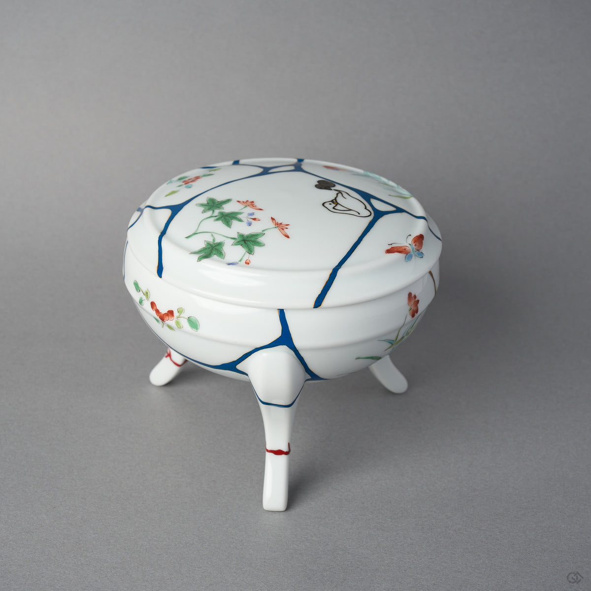 Side view of a kintsugi Arita lidded bowl showing a 24K gold seam descending the body among blue urushi lines, painted blossoms, and vermilion marks on the restored feet.