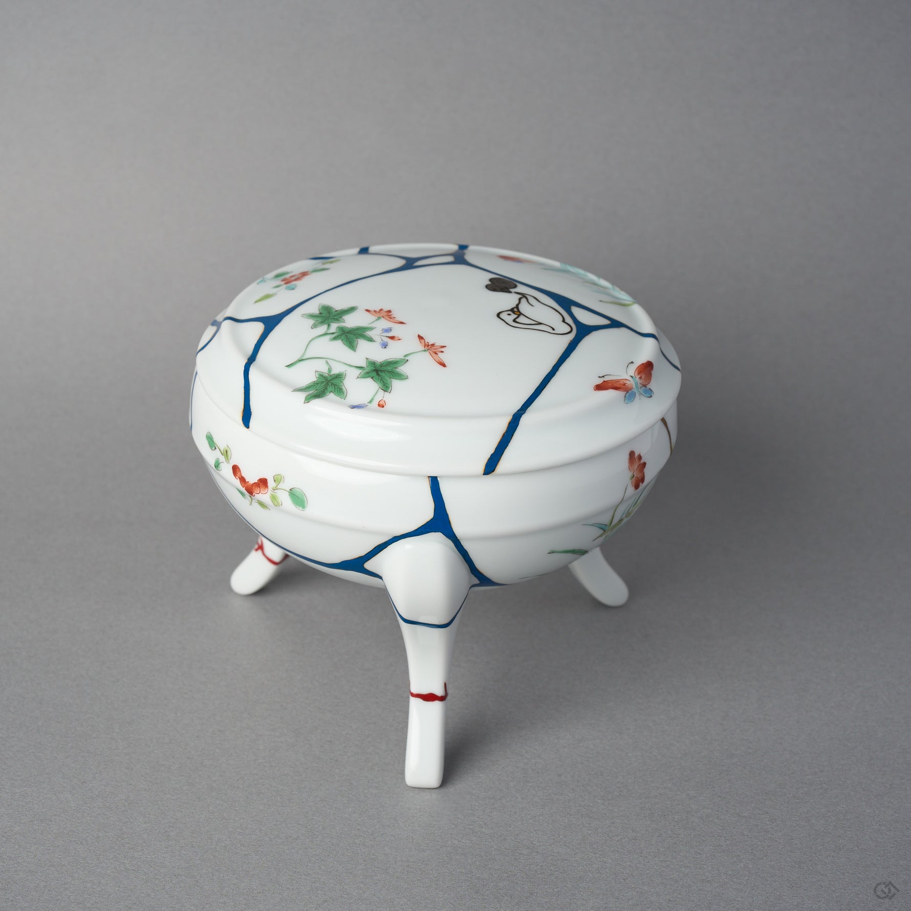 Side view of a kintsugi Arita lidded bowl showing a 24K gold seam descending the body among blue urushi lines, painted blossoms, and vermilion marks on the restored feet.