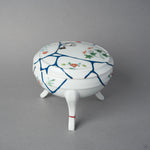 Side view of an Arita porcelain lidded bowl with blue urushi lines crossing the domed lid and body, 24K gold kintsugi finishing, and hand-painted enamel flowers.