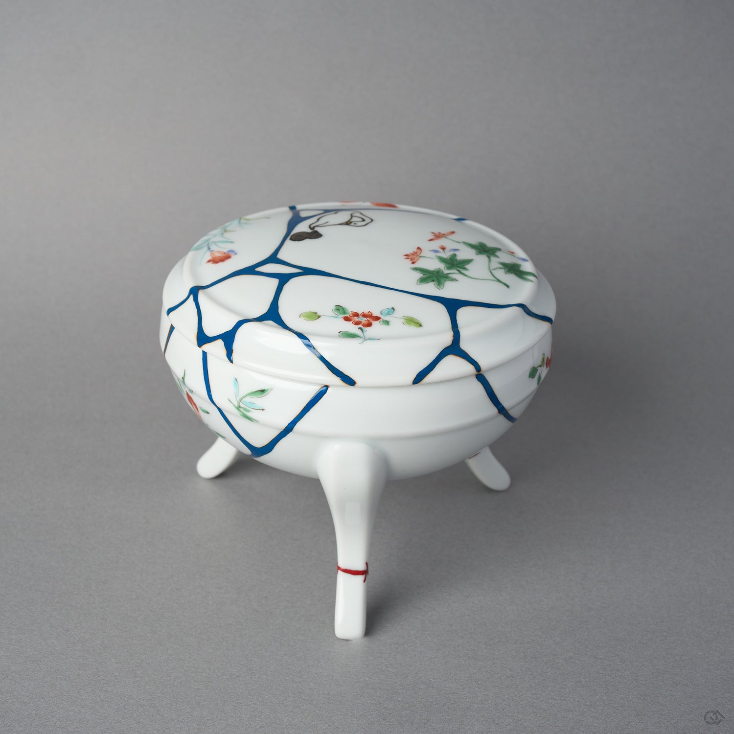 Side view of an Arita porcelain lidded bowl with blue urushi lines crossing the domed lid and body, 24K gold kintsugi finishing, and hand-painted enamel flowers.