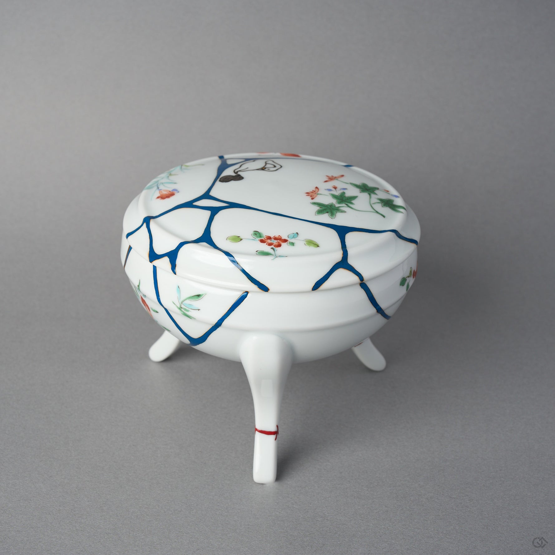 Side view of an Arita porcelain lidded bowl with blue urushi lines crossing the domed lid and body, 24K gold kintsugi finishing, and hand-painted enamel flowers.