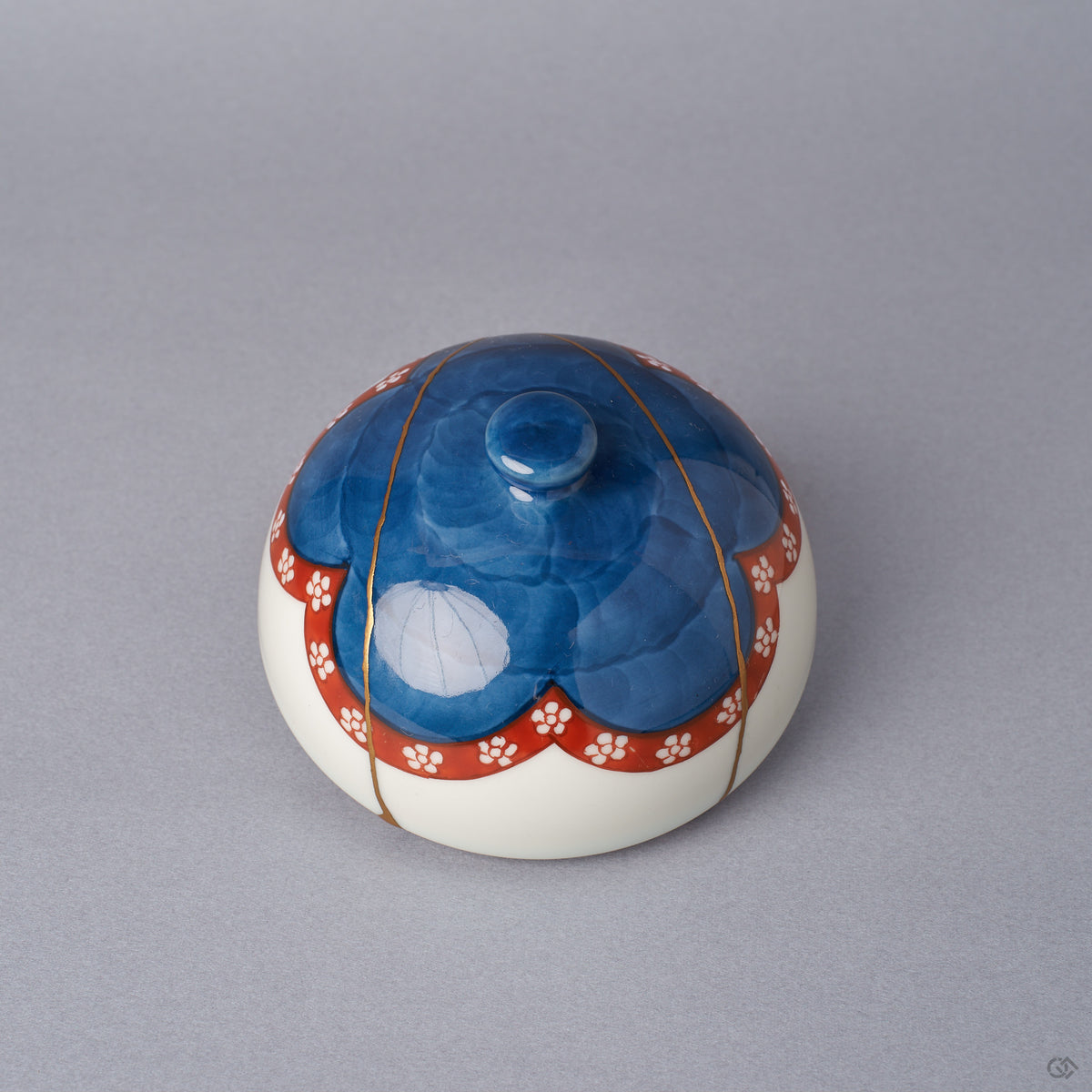 The domed lid of an Arita ware lidded bowl, cobalt-blue crown with a vermilion plum-blossom band, showing 24K gold kintsugi seams tracing the outer surface, viewed from above the side.