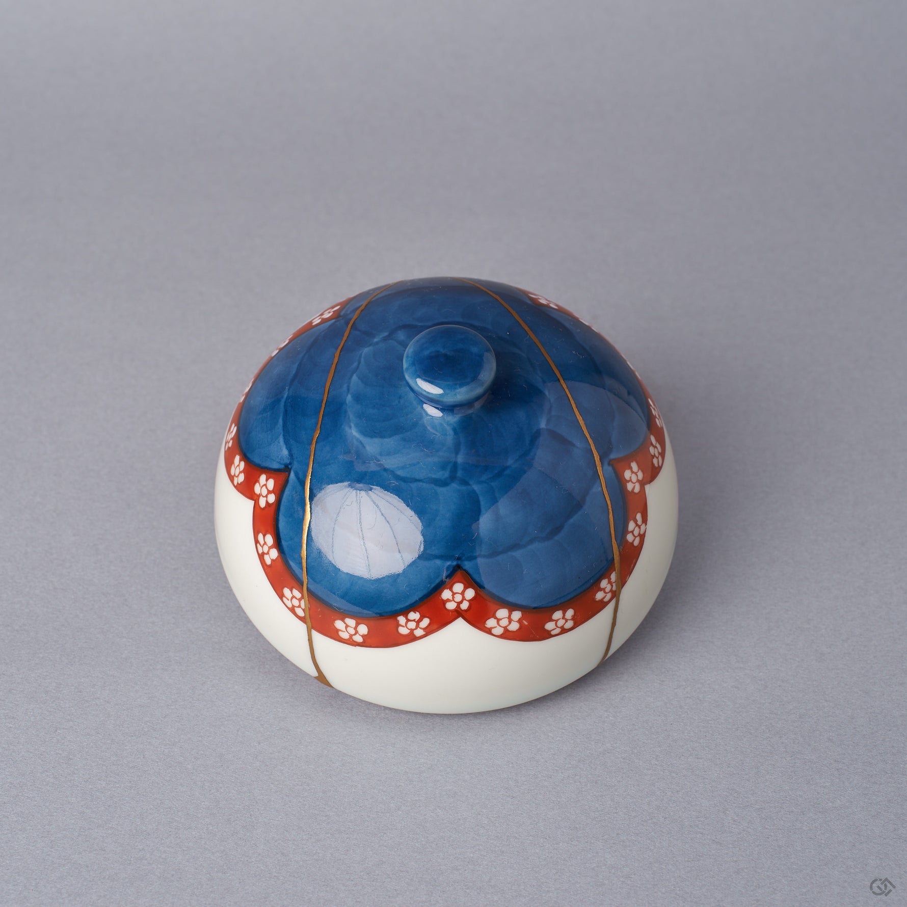 The domed lid of an Arita ware lidded bowl, cobalt-blue crown with a vermilion plum-blossom band, showing 24K gold kintsugi seams tracing the outer surface, viewed from above the side.