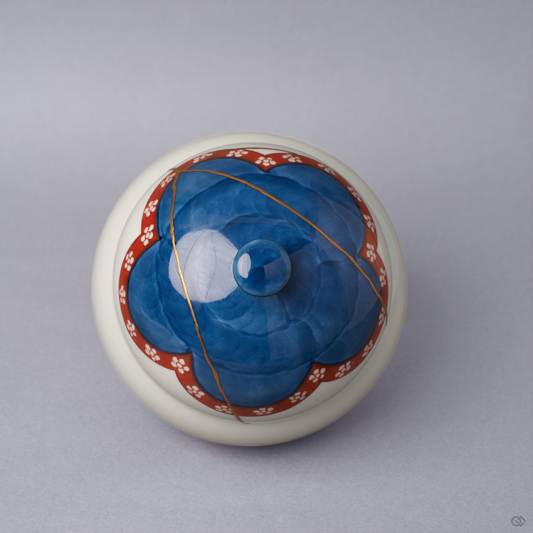 Top view of an Arita ware lidded bowl with the lid in place, a cobalt-blue crown ringed by a vermilion plum-blossom band and a 24K gold kintsugi seam crossing the dome to the knob.