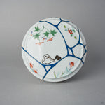 Top view of the Arita lidded bowl lid, blue urushi lines gathered like water across white porcelain, with a swan and cygnet in maki-e among painted blossoms.