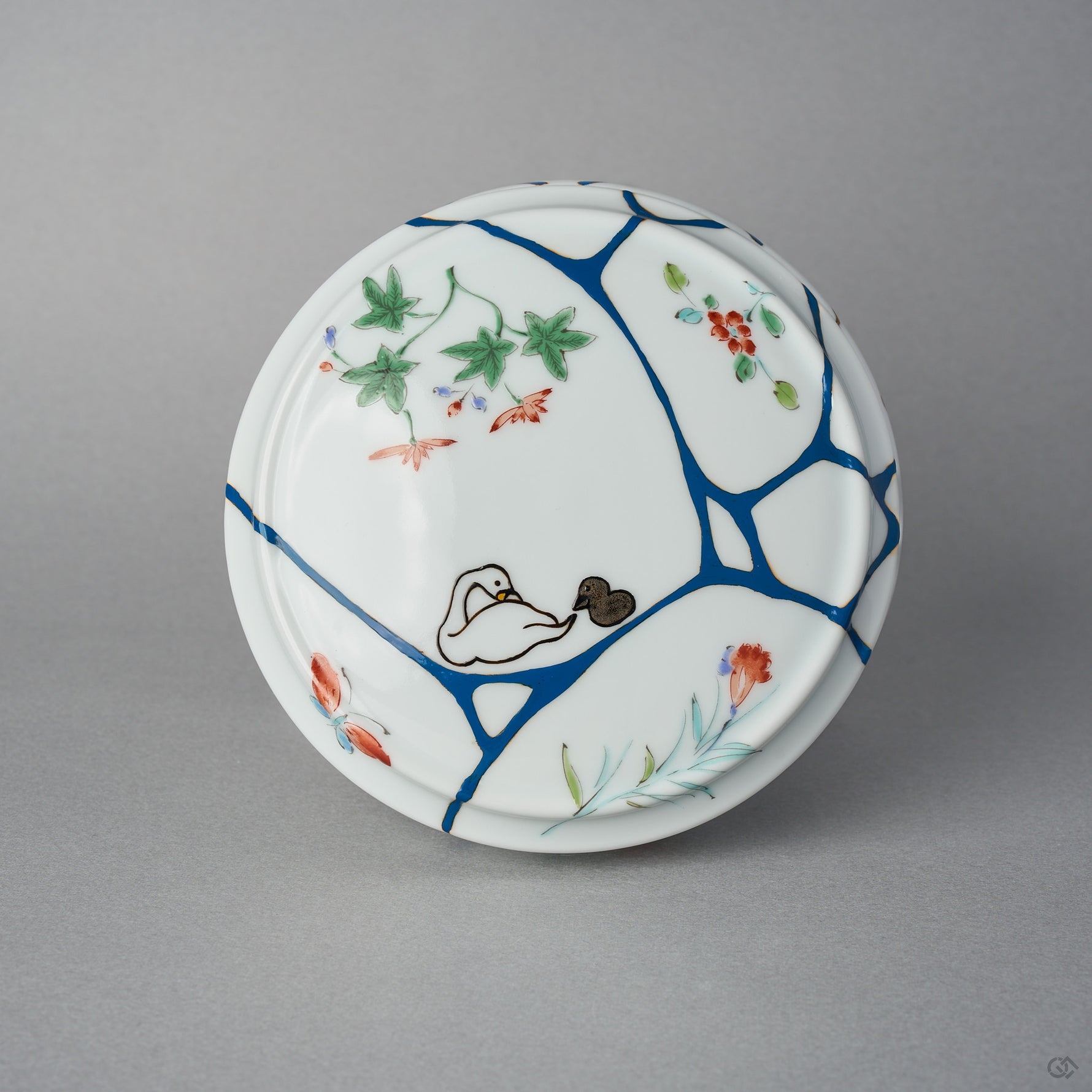 Top view of the Arita lidded bowl lid, blue urushi lines gathered like water across white porcelain, with a swan and cygnet in maki-e among painted blossoms.