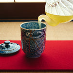 Kintsugi lidded tea cup in Kyoto ware with green tea being poured on a red felt mat, a 24K gold urushi seam on the blue shonzui body.