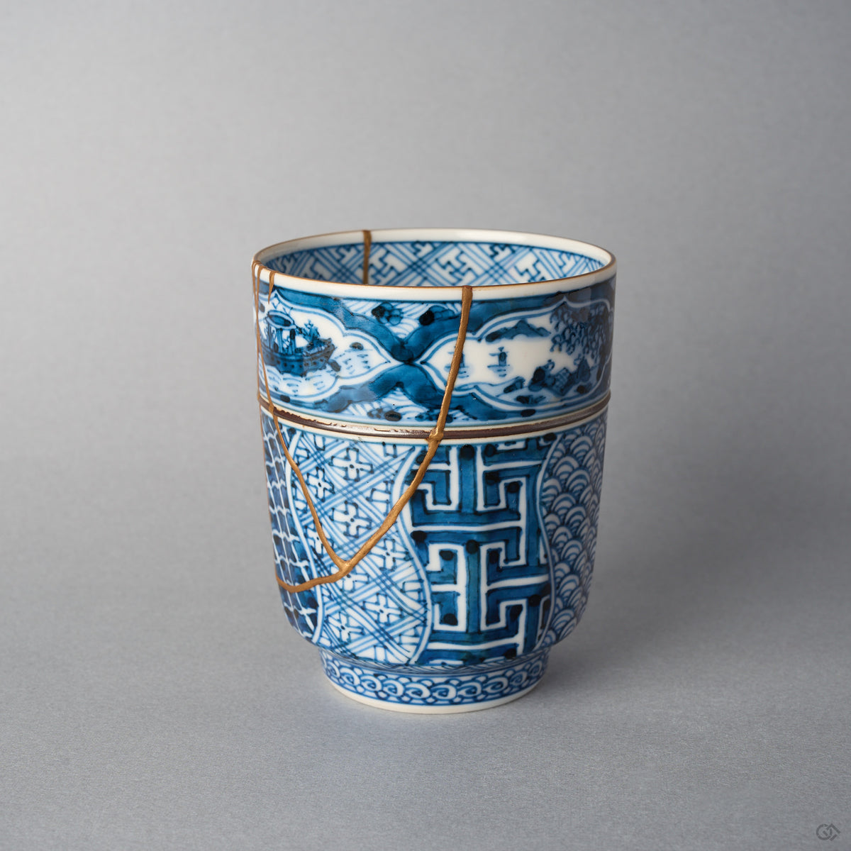 Kintsugi tea cup side view: a gold urushi line follows the painted landscape band and a V-shaped 24K gold seam crosses the shonzui body.