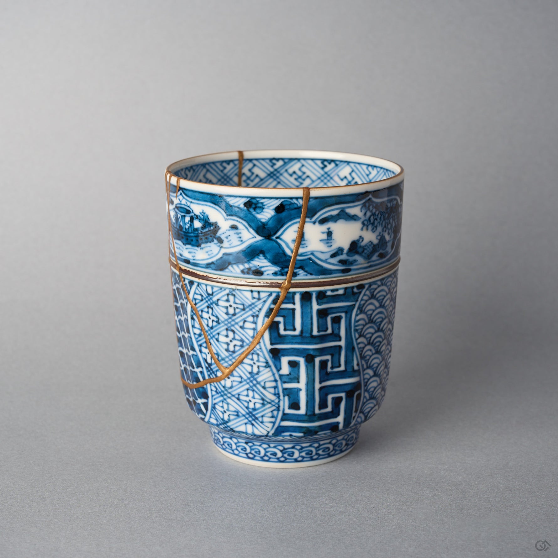 Kintsugi tea cup side view: a gold urushi line follows the painted landscape band and a V-shaped 24K gold seam crosses the shonzui body.