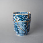 Kintsugi tea cup opposite side: 24K gold urushi seams cross the rim and descend the woven shonzui panels toward the foot.