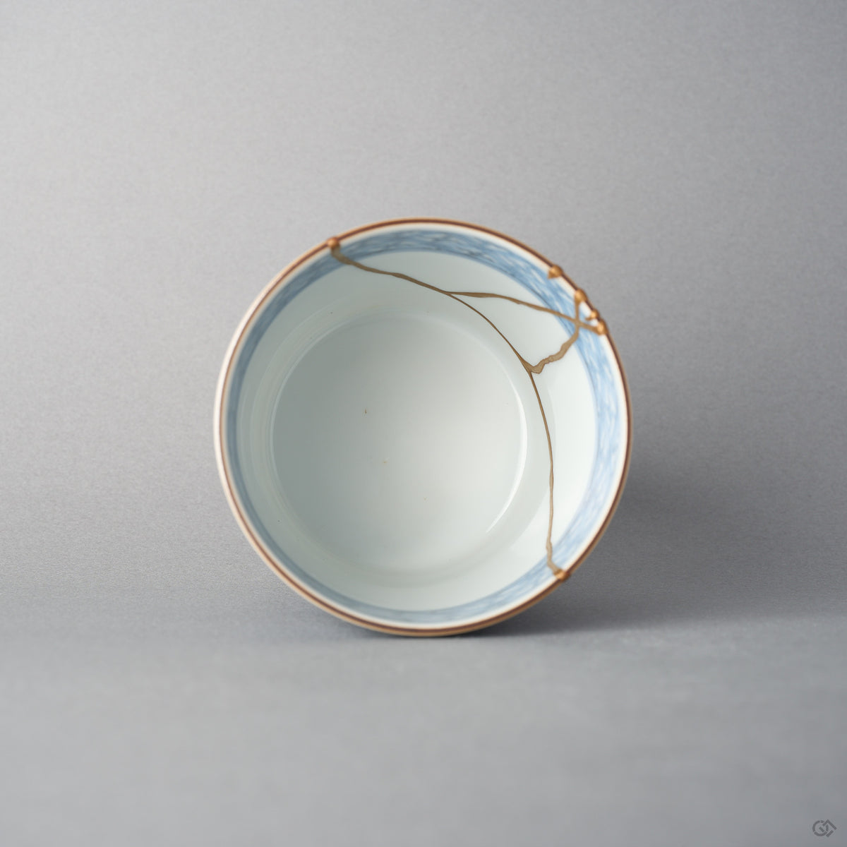 Kintsugi tea cup seen from above; a seam of urushi and 24K gold crosses the blue-banded rim onto the white interior.