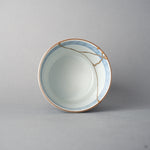 Kintsugi tea cup seen from above; a seam of urushi and 24K gold crosses the blue-banded rim onto the white interior.