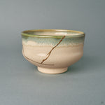 A kintsugi Kyoto Ware matcha bowl seen from the front, its gold seam descending an ivory body beneath a green glaze rim, a quiet beauty.