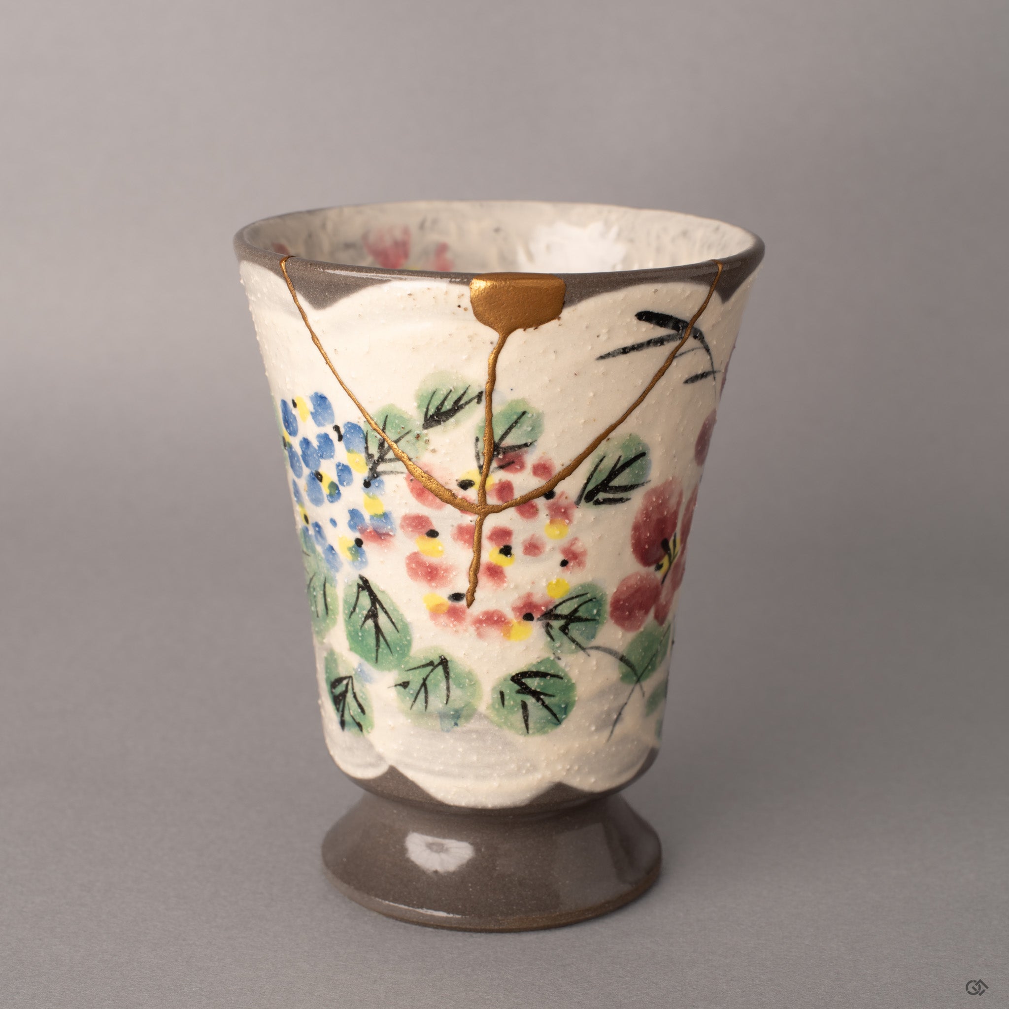 A tall Kyoto mug stands with sculptural presence, gold kintsugi seams threading through hand-painted florals, a philosophy of care made visible.