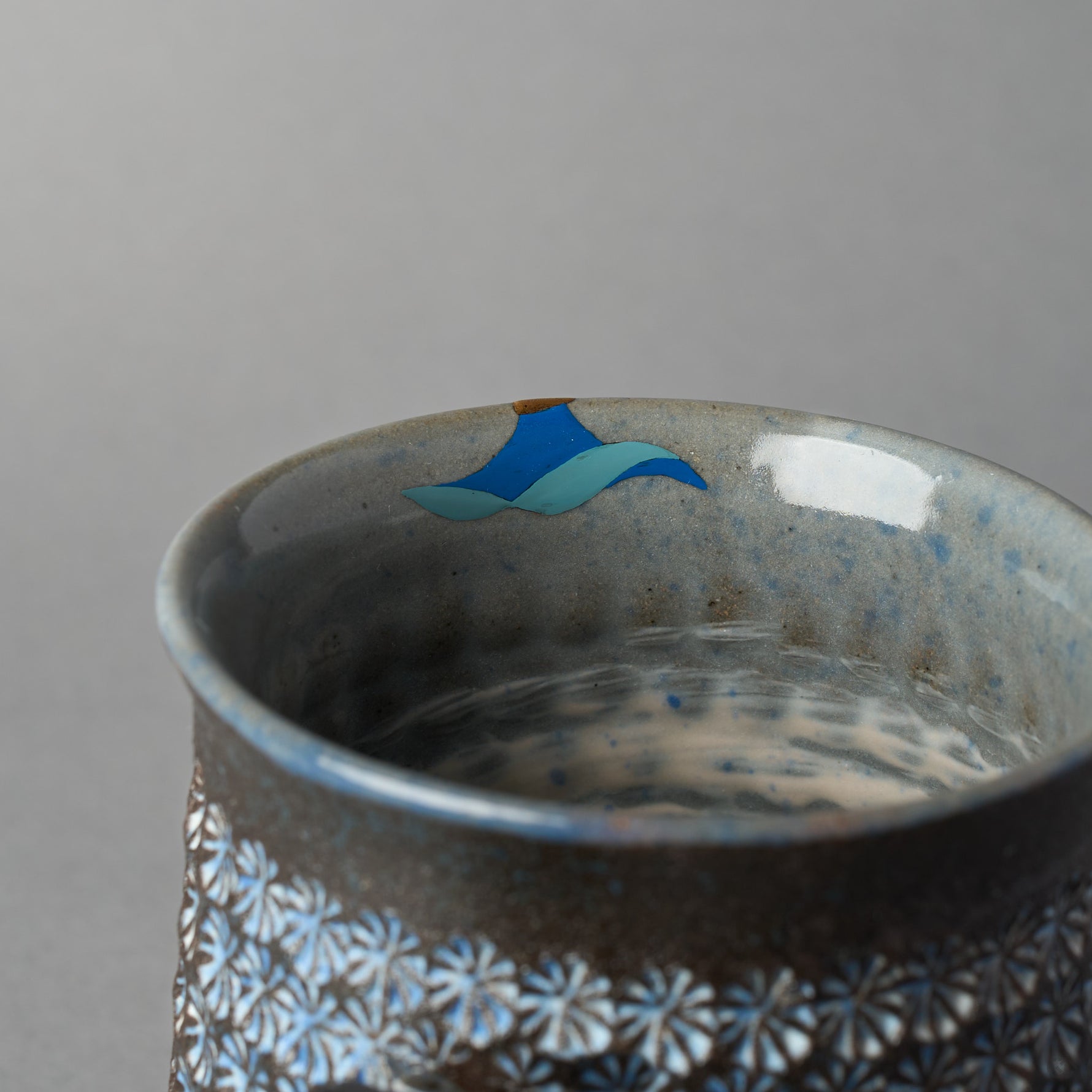 Close-up of the urushi-e painting on the inner rim of the Kyoto ware mug: a rising sun in blue tones beneath the 24K gold kintsugi at the chip, read as sunlight over waves.