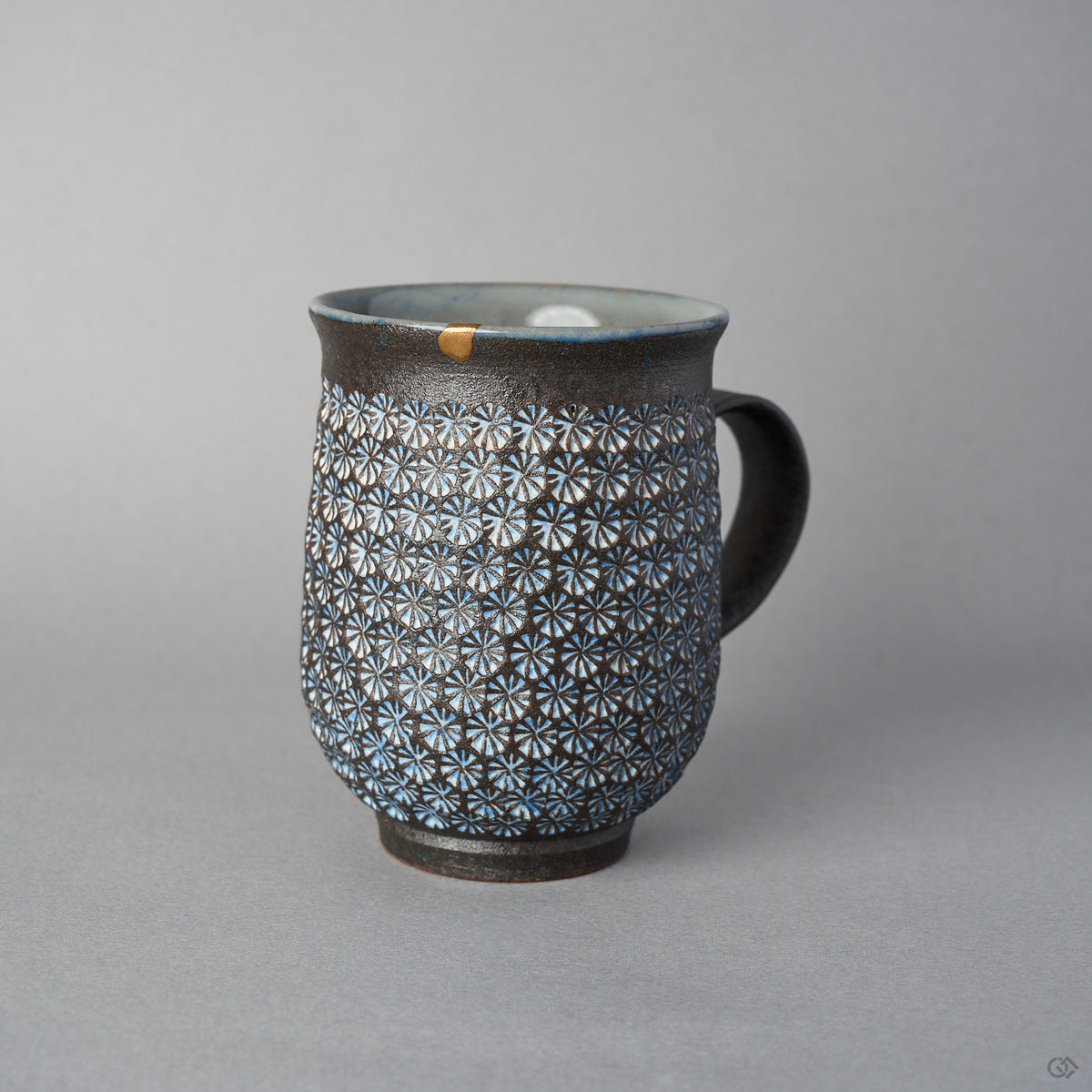 Front view of a Kyoto ware kintsugi mug in dark clay stamped with white mishima florets, a 24K gold rim restoration, flared lip, and curved dark handle.