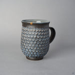 Front view of a Kyoto ware kintsugi mug in dark clay stamped with white mishima florets, a 24K gold rim restoration, flared lip, and curved dark handle.