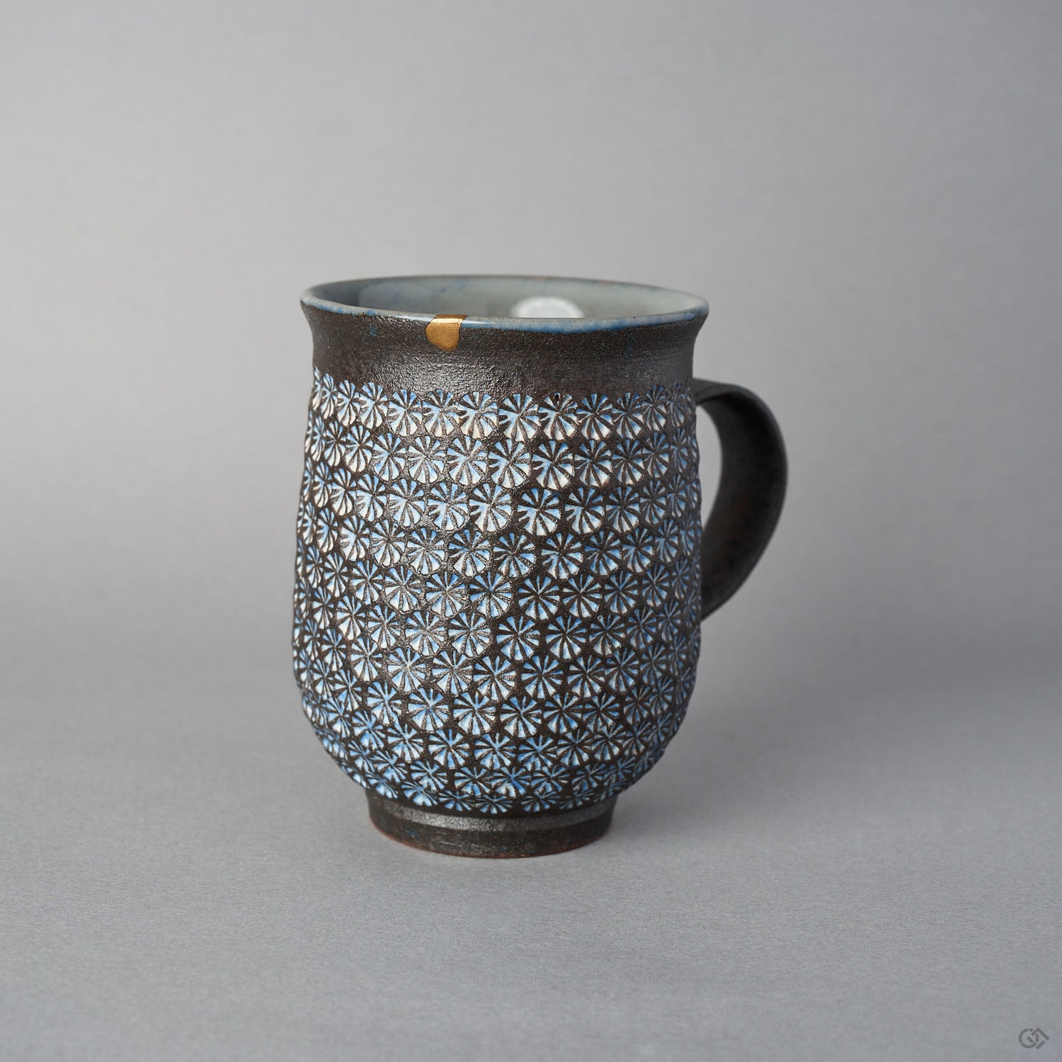 Front view of a Kyoto ware kintsugi mug in dark clay stamped with white mishima florets, a 24K gold rim restoration, flared lip, and curved dark handle.