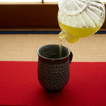Kintsugi Kyoto ware mug on a red felt runner over tatami as green tea is poured, dark mishima-stamped clay with a 24K gold restoration at the rim.