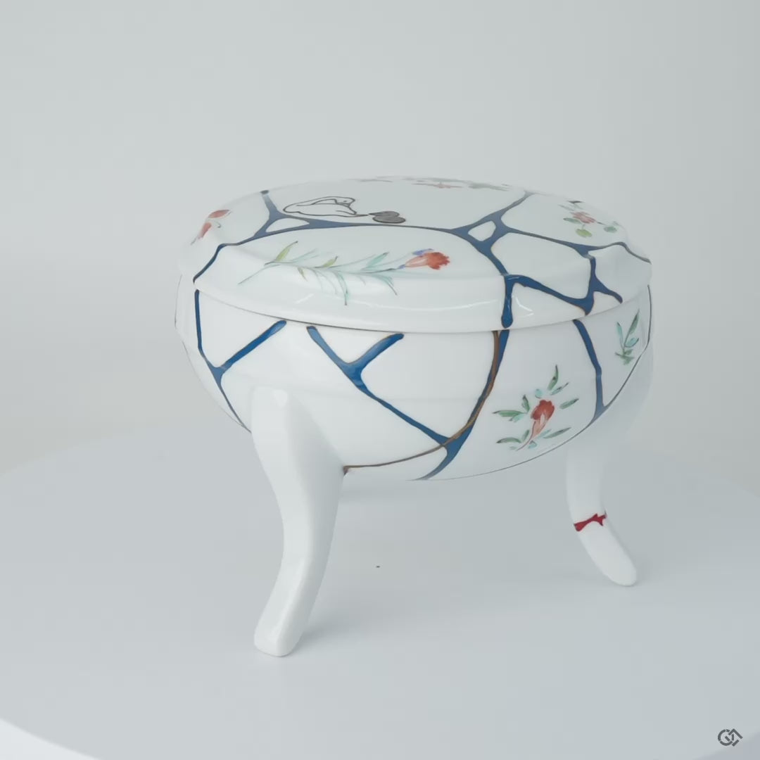 Rotating view of an Arita ware lidded bowl in white porcelain with 24K gold kintsugi seams and blue urushi lines, turning to show the full restoration.