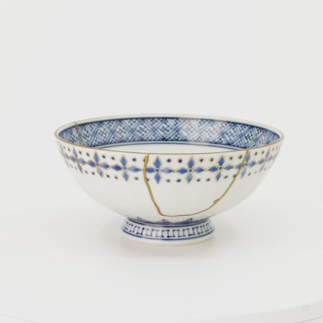 A 360-degree turn of the kintsugi rice bowl, gold seams and blue diamonds revolving slowly into view, scars as beauty seen from every side.