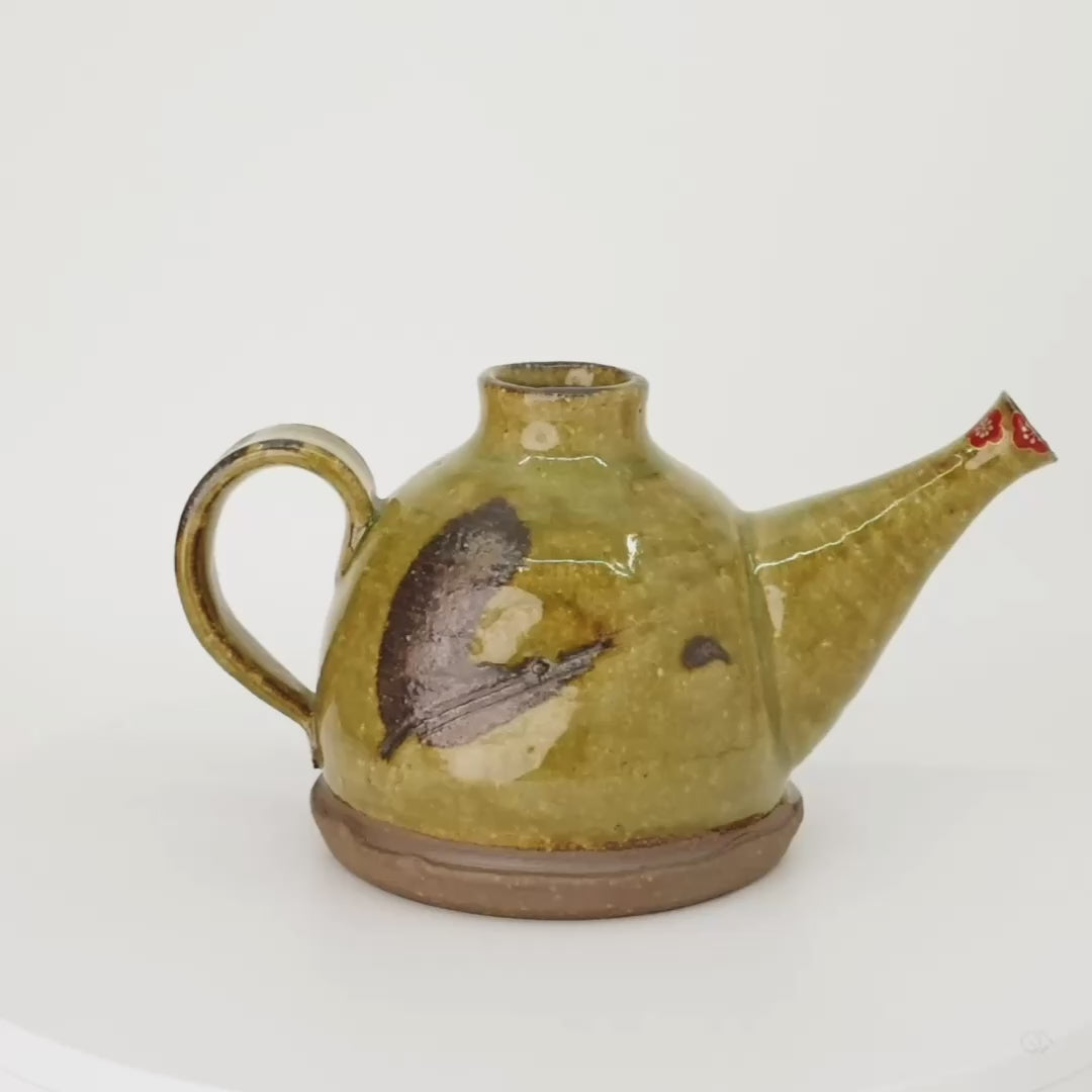 Rotation video of a Kyoto ware earthenware sake carafe, ochre Oribe-green crackle glaze and iron-glaze brushwork, with a gold-and-silver plum blossom restored at the spout.
