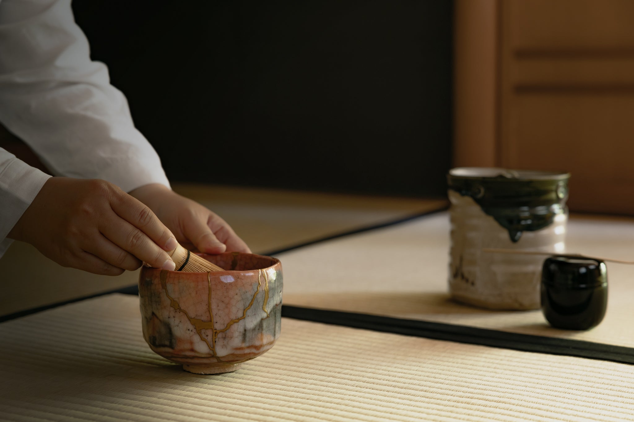 Whisking matcha in a Kintsugi-restored Raku-style bowl inside a Japanese tea room, accompanied by chasen, natsume, and traditional tea ceremony utensils.