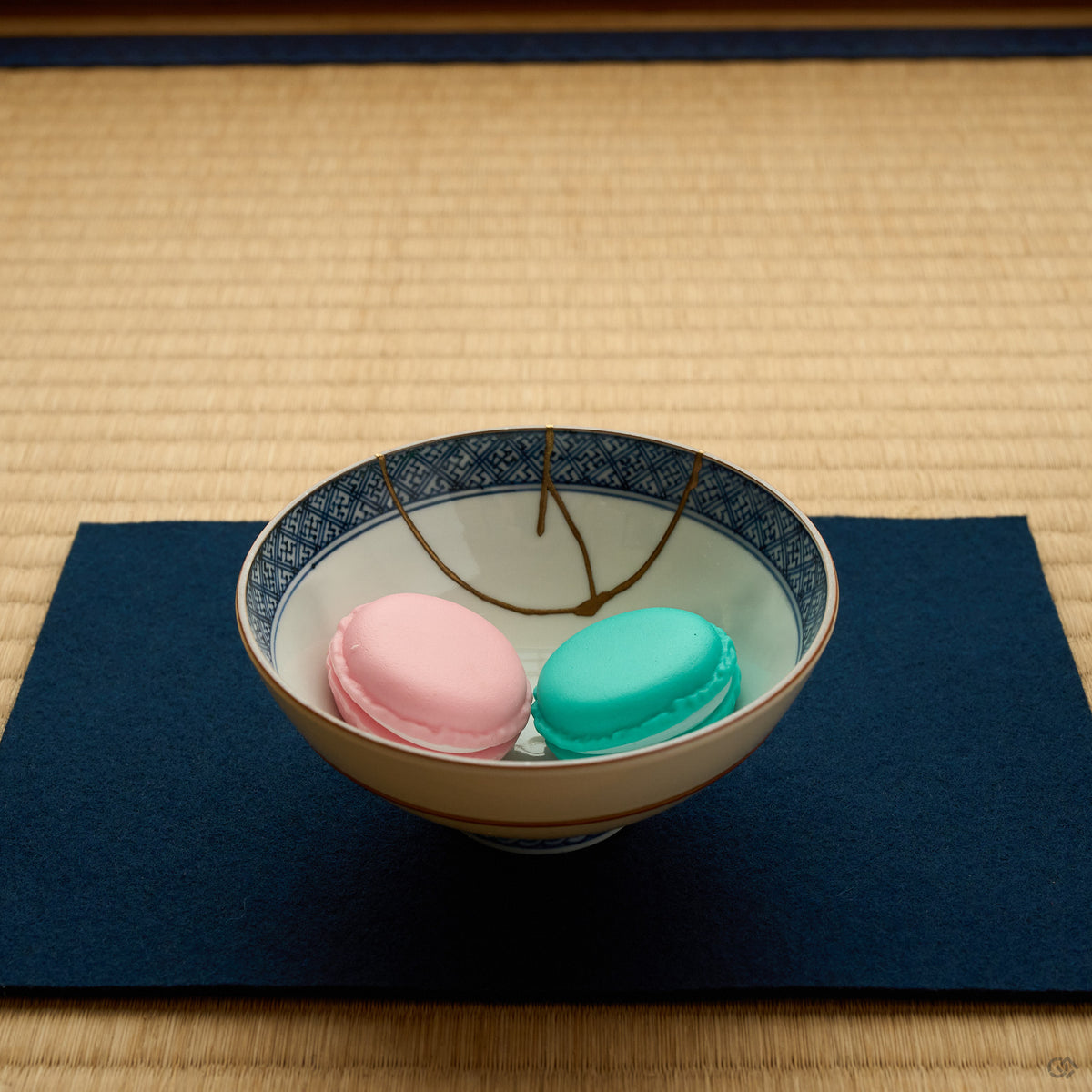 Kintsugi Kyoto ware rice bowl on an indigo cloth over tatami, holding pink and green macarons, the 24K gold seam crossing its cobalt lattice rim.