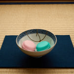 Kintsugi Kyoto ware rice bowl on an indigo cloth over tatami, holding pink and green macarons, the 24K gold seam crossing its cobalt lattice rim.
