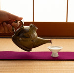 Kyoto ware kintsugi sake carafe pouring into a white sake cup on a tatami setting, ochre Oribe-green crackle glaze, gold-and-silver plum blossom at the spout.