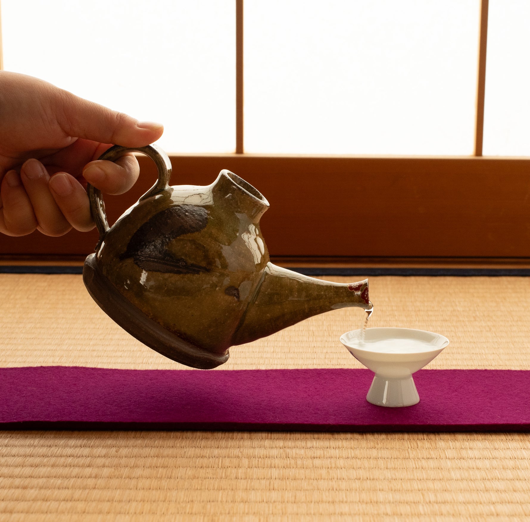 Kyoto ware kintsugi sake carafe pouring into a white sake cup on a tatami setting, ochre Oribe-green crackle glaze, gold-and-silver plum blossom at the spout.