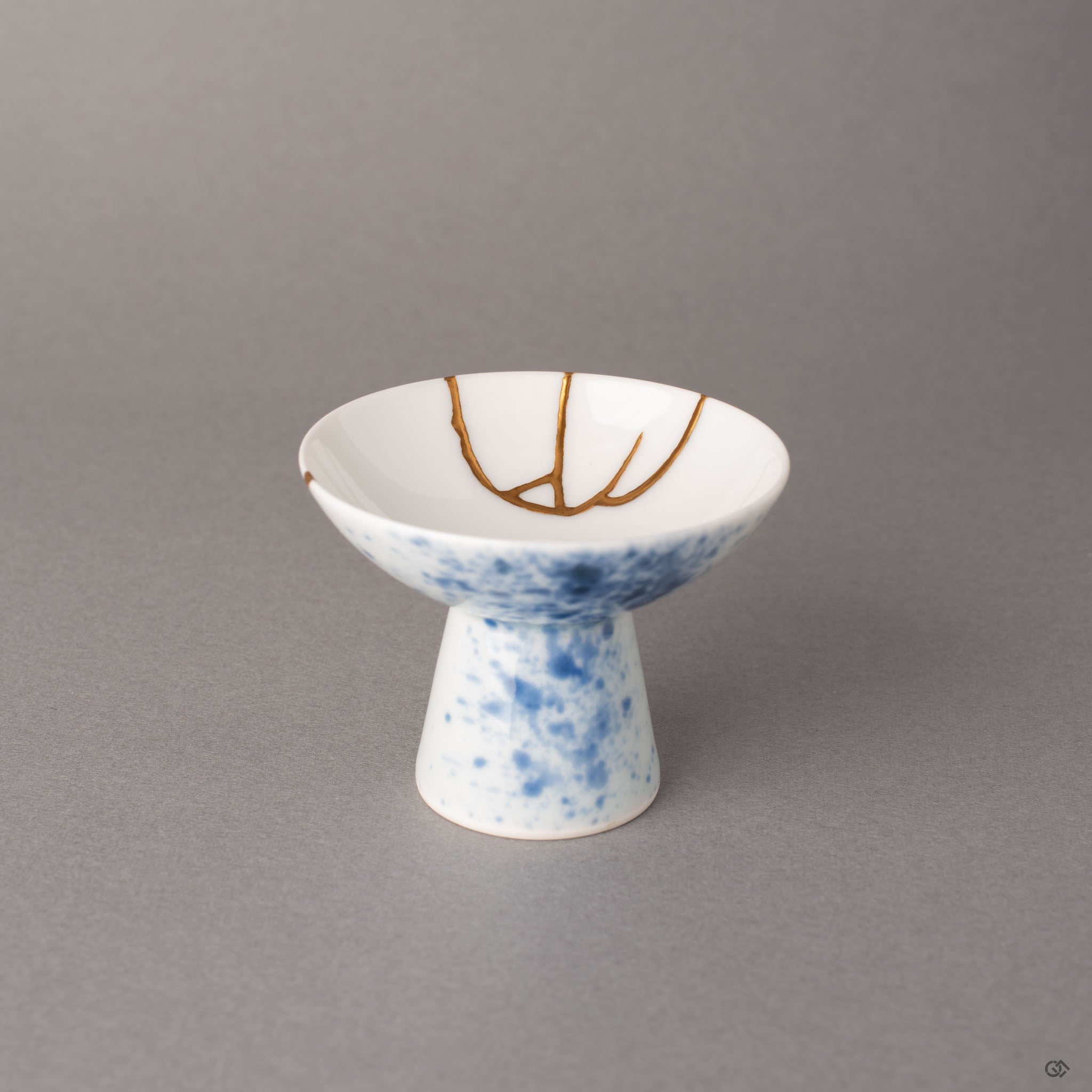 Kintsugi Arita sake cup with branching gold seams across white porcelain and sometsuke blue splatter, a quiet presence on a sculptural pedestal.