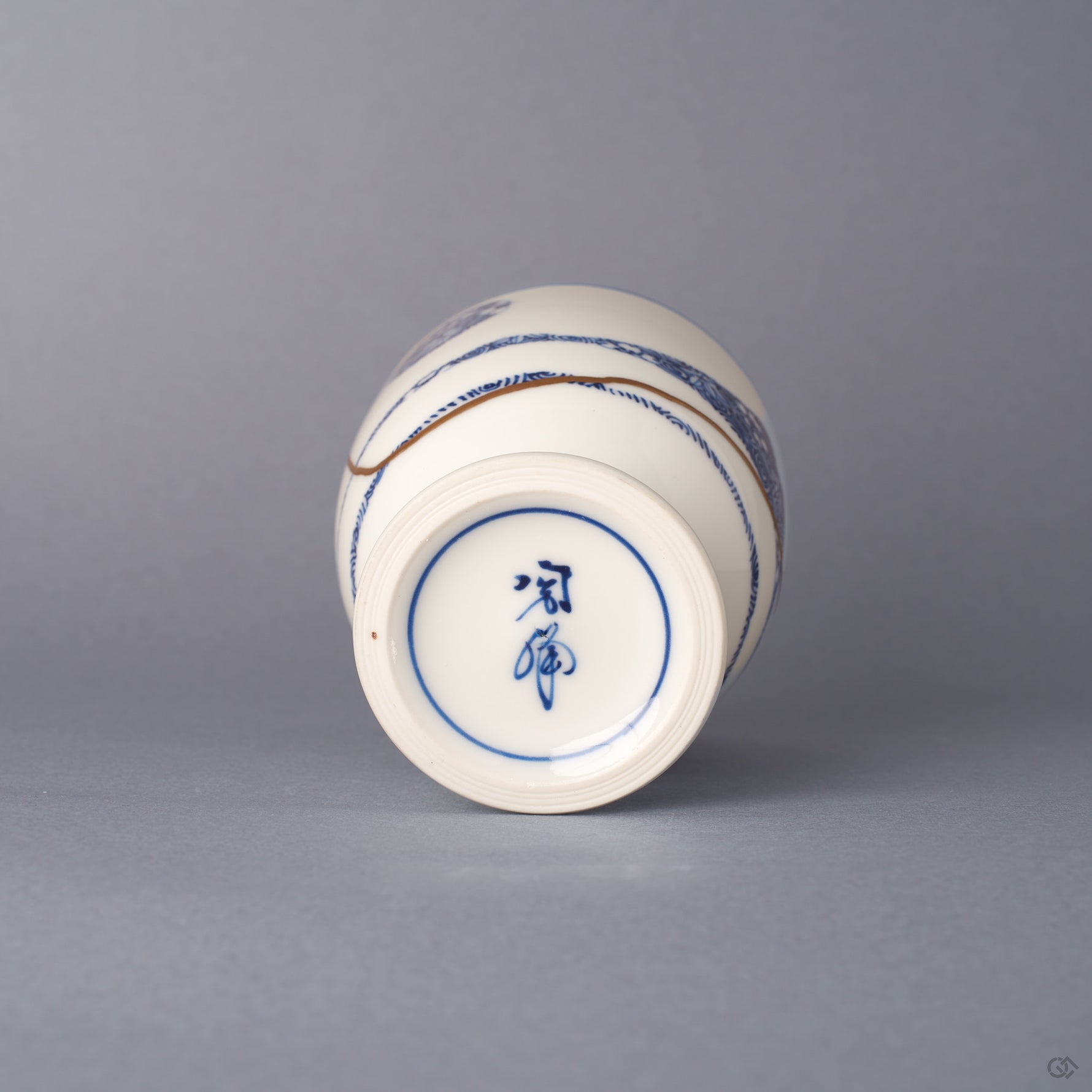 Underside of the Arita ware sake cup in white porcelain, showing the turned foot, a hand-painted cobalt maker's mark, and 24K gold kintsugi seams reaching toward the base.
