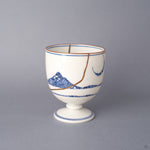 Front view of an Arita ware sake cup in white porcelain, cobalt-blue sometsuke bands and a floral roundel, with 24K gold kintsugi seams crossing the rounded body.