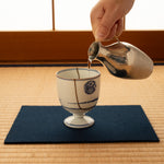 Arita ware sake cup in white porcelain with cobalt-blue bands and 24K gold kintsugi seams, being filled with sake poured from a hammered silver server in a tatami setting.