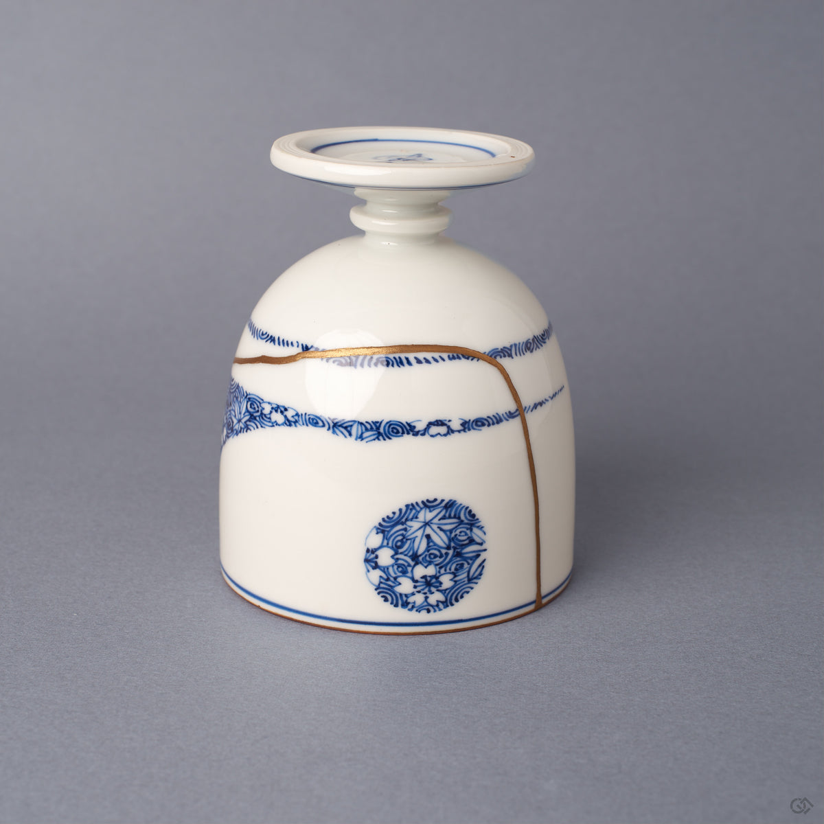 Side view of a footed Arita ware sake cup in white porcelain, its 24K gold kintsugi seams crossing cobalt-blue patterned bands around the body.