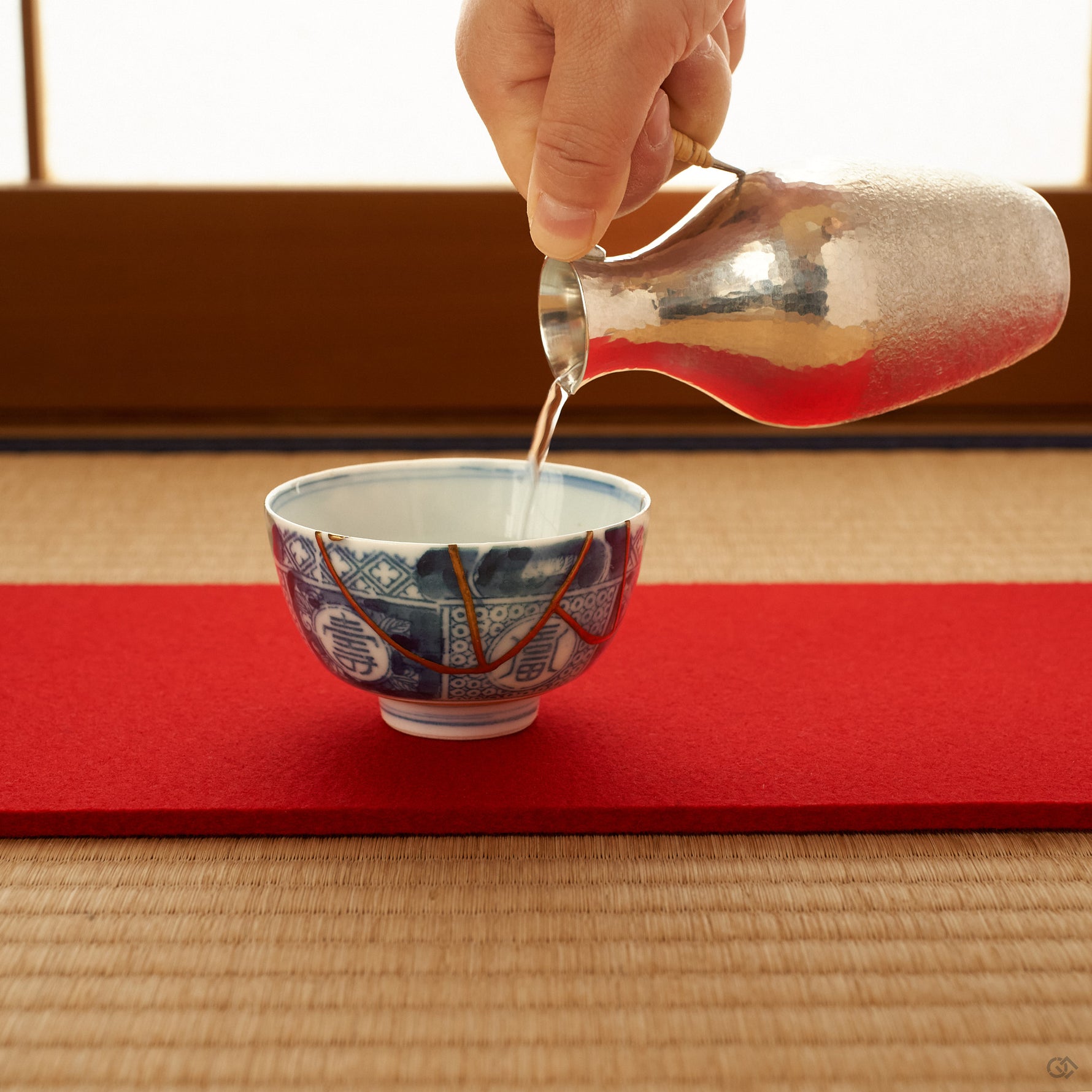 Blue and white Ko-Imari sake cup with 24K gold kintsugi seams on a red felt runner over tatami, receiving sake poured from a hammered silver carafe in soft window light.