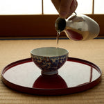 Sake poured from a hammered silver carafe into a kintsugi Ko-Imari cup on a red lacquer tray over tatami, gold arcs crossing its blue karakusa vines.