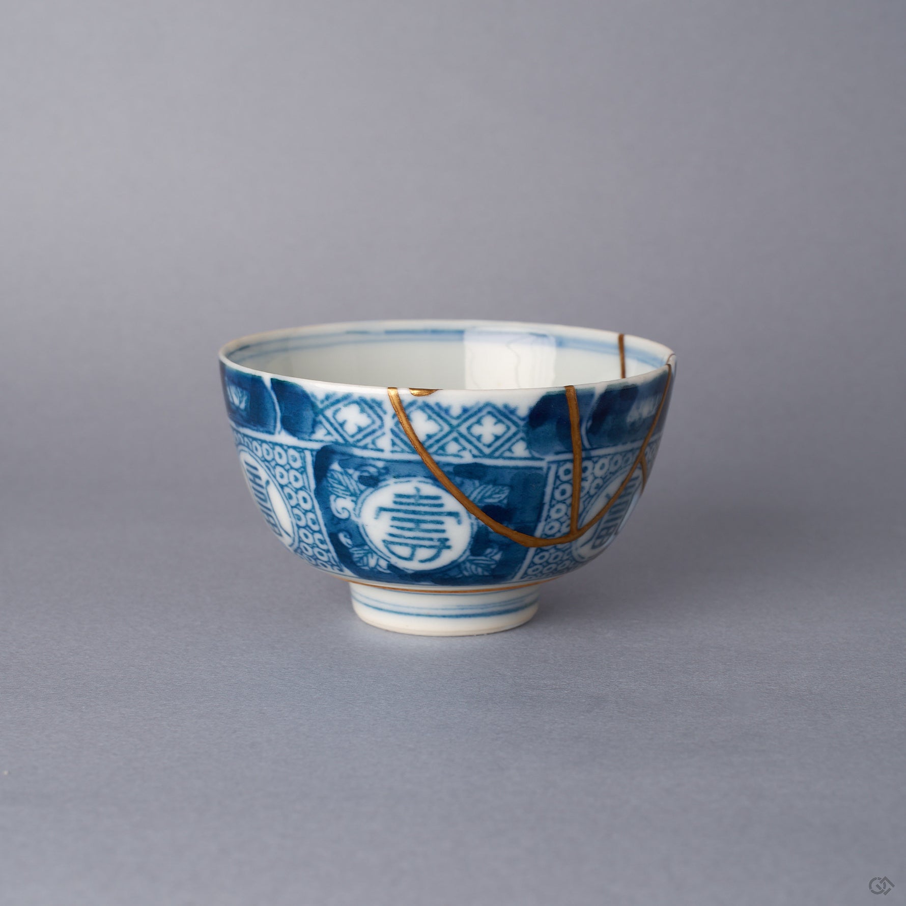 Side view of a Ko-Imari sake cup in sometsuke blue and white porcelain, a 24K gold kintsugi seam curving from the rim past a round longevity medallion toward the foot.