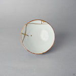 Top view of a kintsugi Ko-Imari sake cup, the white interior crossed by 24K gold restoration lines with a single gold drop resting near the well.