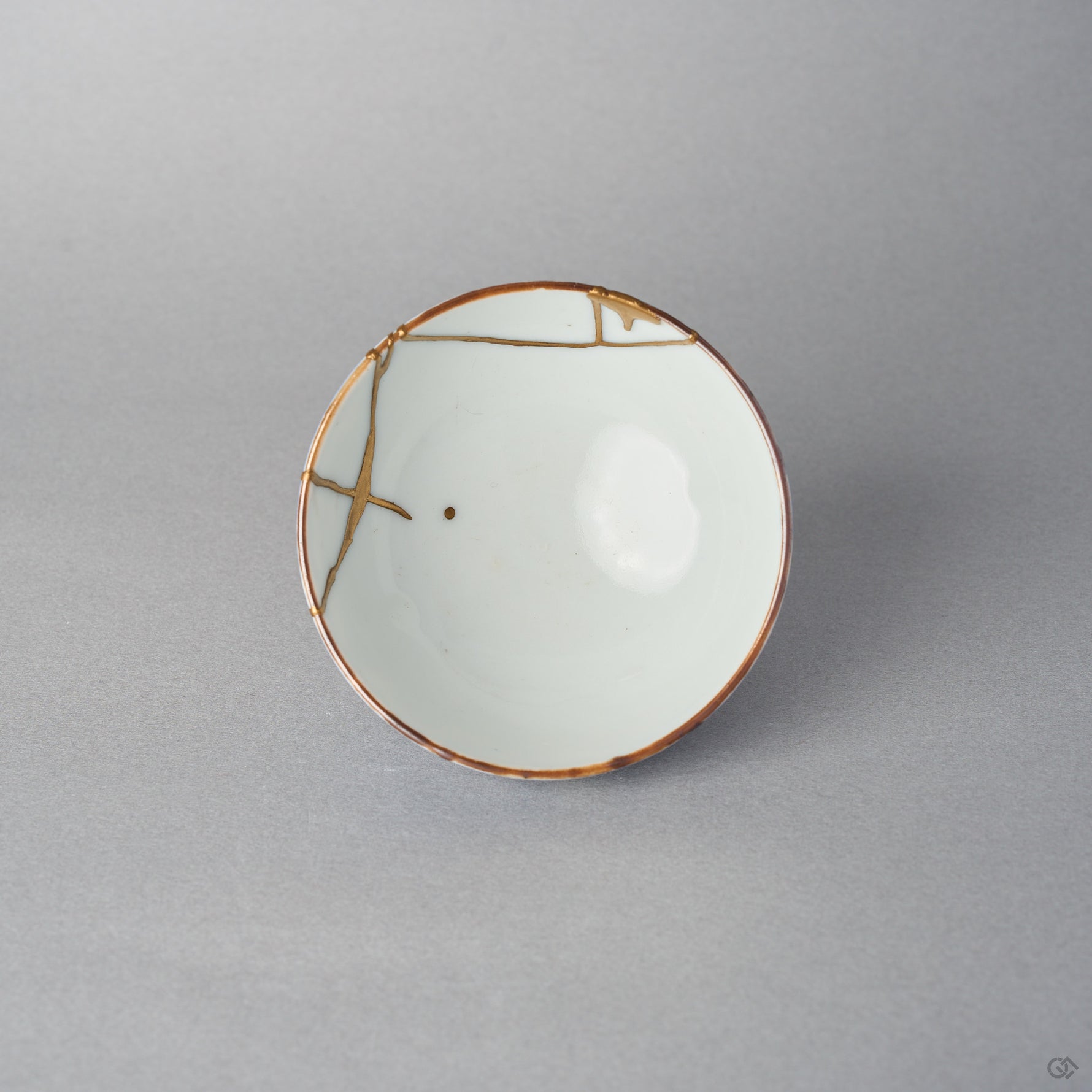 Top view of a kintsugi Ko-Imari sake cup, the white interior crossed by 24K gold restoration lines with a single gold drop resting near the well.