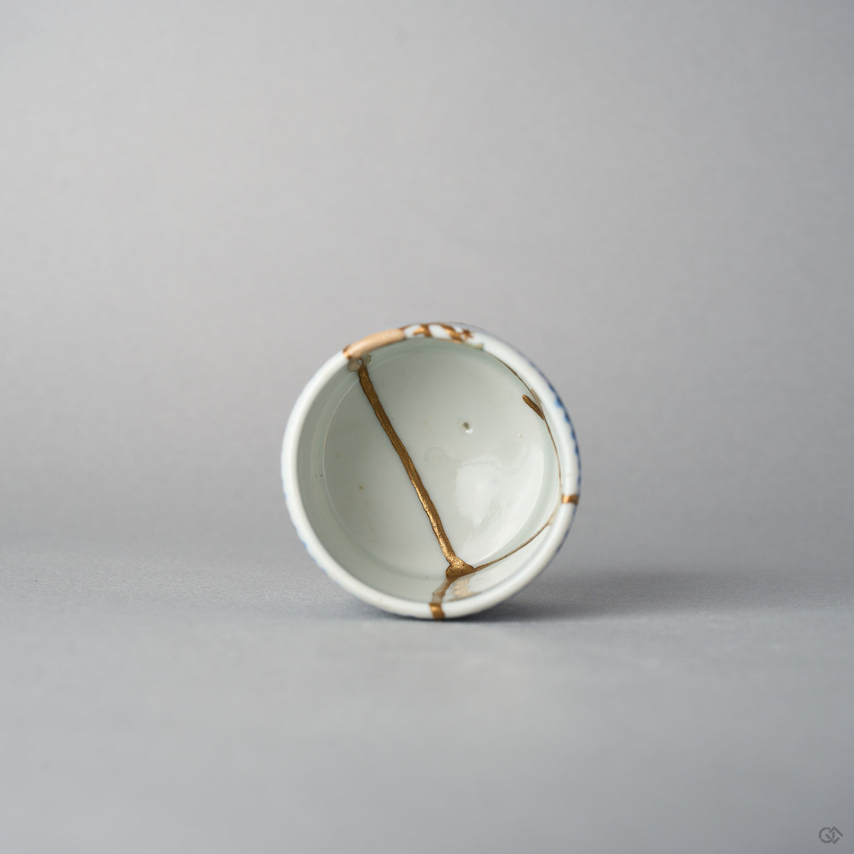 Top view of a Ko-Imari sake cup showing 24K gold kintsugi lines converging inside the white well, the rim and outer wall patterned in sometsuke blue takokarakusa, urushi-restored.