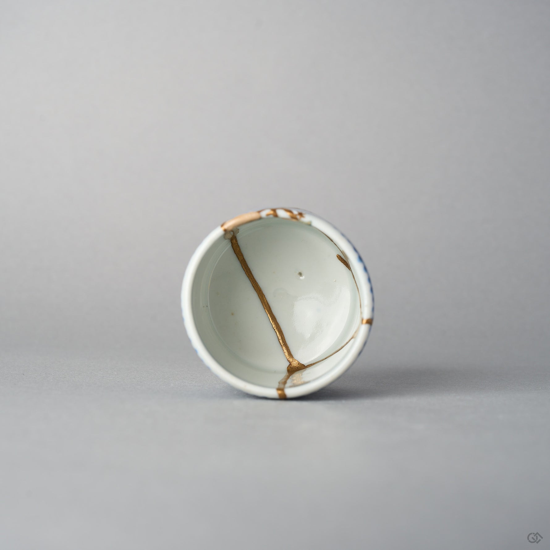 Top view of a Ko-Imari sake cup showing 24K gold kintsugi lines converging inside the white well, the rim and outer wall patterned in sometsuke blue takokarakusa, urushi-restored.