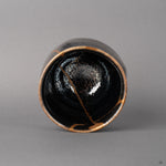 From above, the tenmoku cup reveals scattered silver flecks and kintsugi lines descending inward, a quiet beauty in the round.