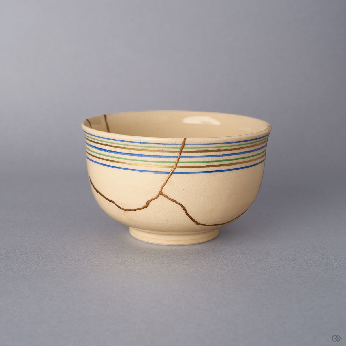 Front view of a Kyoto ware kintsugi cup in cream colored earthenware, with blue, green, and gold koma-suji rim lines and a network of seams restored in 24K gold.