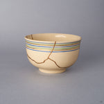 Front view of a Kyoto ware kintsugi cup in cream colored earthenware, with blue, green, and gold koma-suji rim lines and a network of seams restored in 24K gold.