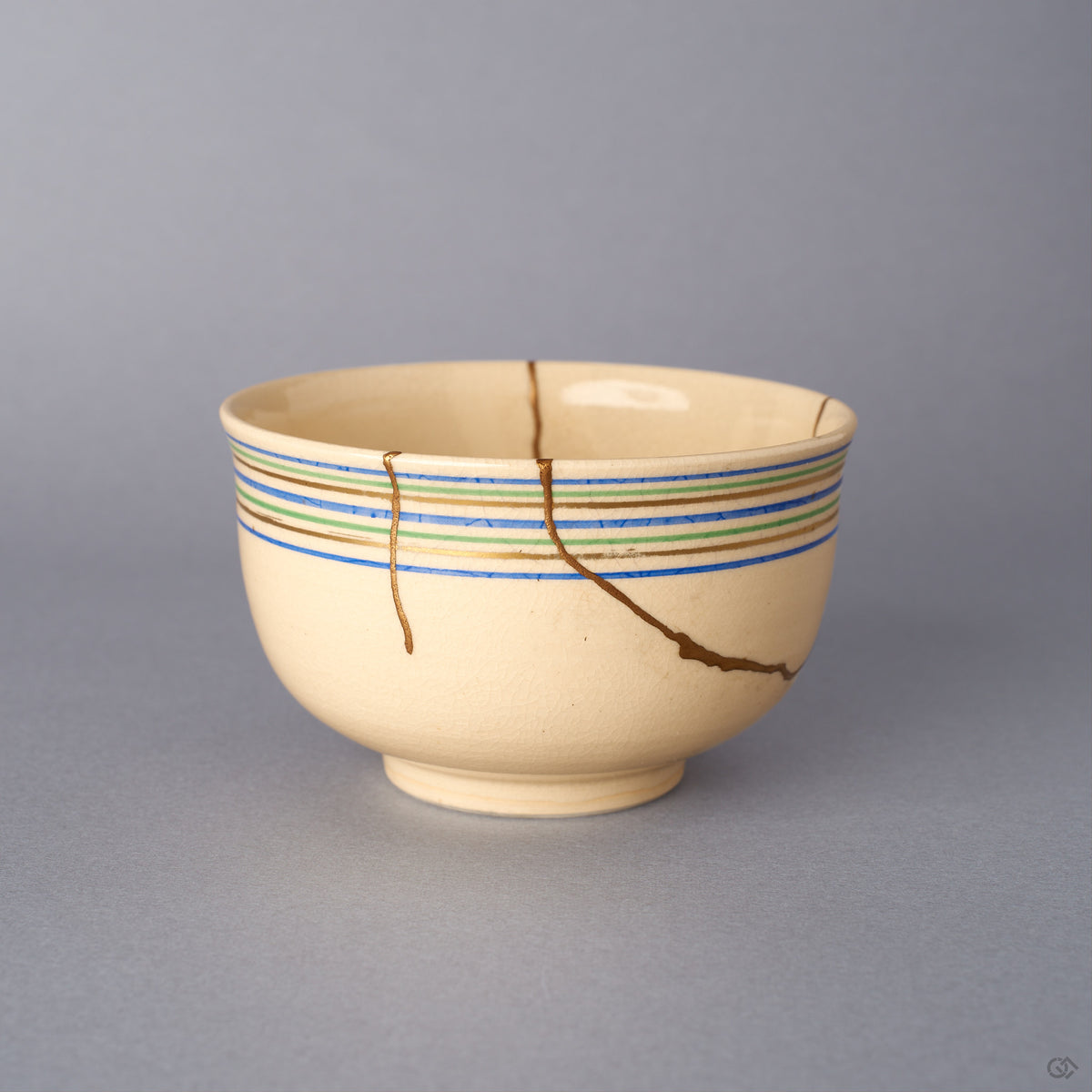 Opposite side view of the Kyoto ware kintsugi cup in cream earthenware, where 24K gold seams branch across the lower body below the blue, green, and gold rim lines.